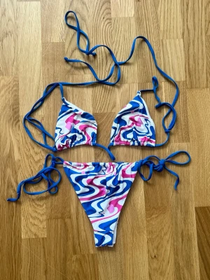 Färgglad bikini med blå snörning - Snygg bikini med trendigt vågigt mönster i blått, rosa och vitt. Klassisk trekantsmodell med knyt i sidorna och runt nacken. Perfekt för sommaren och strandhäng. Materialet känns mjukt och följsamt mot huden.