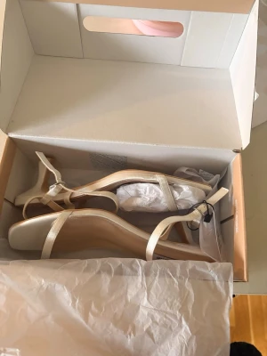 Krämvita sandaler från  - Säljer ett par helt oanvända  stilrena krämvita sandaler  med smala remmar och fyrkantig tå. Sandalerna har en låg klack och justerbart spänne vid ankeln. Perfekta till sommarens outfits och lätta att matcha med olika stilar.