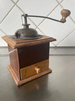 Retro kaffekvarn i trä och metall - Säljer en klassisk manuell kaffekvarn i brunt trä med vev och lock i metall. Kvarnen har en utdragbar låda i ljust trä för det malda kaffet och en snygg träknopp på veven. Perfekt för dig som gillar vintage köksprylar.