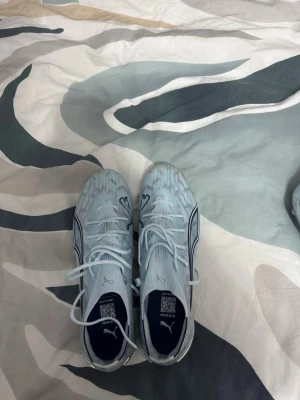 Ljusblåa Puma Ultra fotbollsskor - Säljer ett par ljusblåa Puma Ultra fotbollsskor med svarta detaljer och snörning. Skorna har en modern, strömlinjeformad design och är tillverkade i syntetmaterial för lätt vikt och bra bollkontroll. Perfekta för dig som vill ha stil och prestanda på planen.