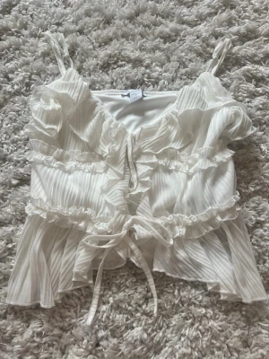 Vit volangig linnetopp från Amisu - Supersöt vit linnetopp från Amisu med tunna axelband, volangdetaljer och knyt framtill. Tunt, plisserat tyg som ger en luftig känsla och romantisk look. Perfekt att styla med jeans eller kjol för en trendig outfit.