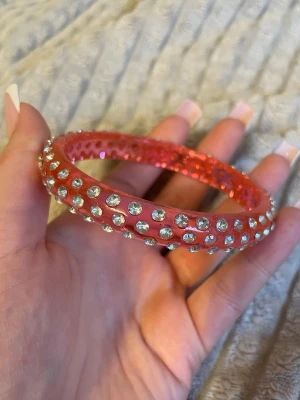 Rosa armband med strassdetaljer - Snyggt armband i genomskinlig rosa plast med massor av glittrande strass-stenar runt hela bandet. Armbandet har en rund form och ger en riktigt poppig och trendig känsla. Perfekt för dig som vill sticka ut med accessoarerna och älskar färg och bling.