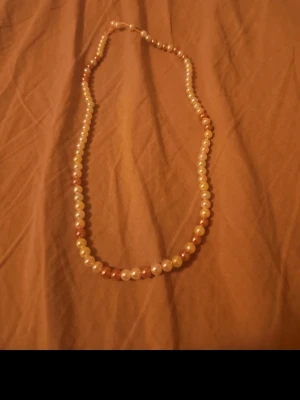 Flerfärgat pärlhalsband - Säljer ett pärlhalsband med runda pärlor i olika färger som vitt, gult och brunt. Halsbandet har en klassisk design och pärlorna är jämnt fördelade längs hela kedjan. Perfekt för dig som gillar en tidlös och elegant stil.