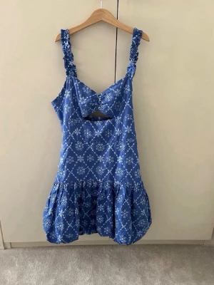 Blå kortklänning med volang och cut-out - Supersöt blå kortklänning med vita blommönster, volangig kjol och trendig cut-out framtill. Klänningen har breda, rynkade axelband och smock i ryggen för skön passform. Perfekt för sommarens alla tillfällen!