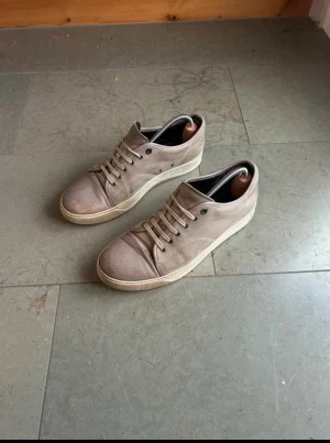Beigea Lanvins  - Stilrena beige sneakers i mocka med vita gummisulor och matchande beige skosnören. Skorna har en klassisk låg profil och diskreta metalldetaljer vid snörningen. Perfekta för en clean och avslappnad look.