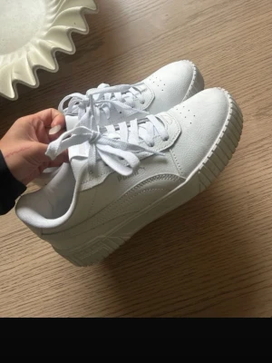 Vita Puma sneakers i läder - Stilrena vita sneakers från Puma i läder med klassisk låg siluett. Skorna har perforerade detaljer på tån, vita skosnören och en chunky sula som ger en modern vibe. Perfekta för dig som vill ha en clean och sportig look.