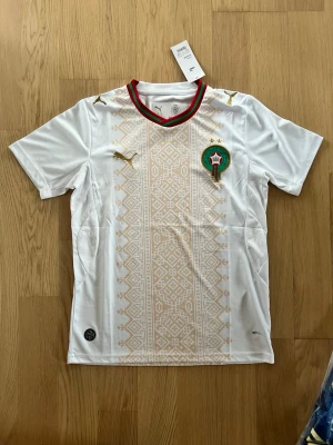 Marocko fotbollströja VM 2026 kit  - Vit fotbollströja från Puma med Marockos landslagsemblem på bröstet, guldfärgade Puma-loggor och ett mönster i beige längs framsidan. Kragen har röda och gröna detaljer. Perfekt för dig som vill stötta Marockos landslag.