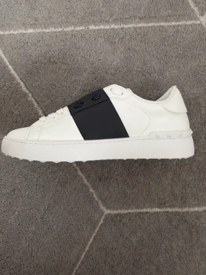 Valentino vita sneakers med navy - Snygga vita sneakers från Valentino med en bred svart läderdetalj över snörningen. Skorna har klassisk låg profil, vita skosnören och diskret logga på plösen. Tillverkade i slätt läder med en chunky vit sula som ger en modern look.