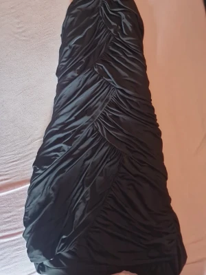 Svart omlott kortklänning i satin - Säljer en svart kort klänning i glansigt satinmaterial med omlottdetaljer och draperad design. Klänningen är axelbandslös och har en snygg, figurnära passform som ger en elegant look. Perfekt för dig som vill sticka ut med en unik stil. Pris kan diskuteras🫶🏼