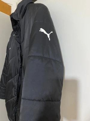 Svart pufferjacka från Puma - Säljer en svart pufferjacka från Puma med klassisk logga på ärmen. Jackan har hög krage och breda, vadderade paneler som håller dig varm. Perfekt för kalla dagar och har en sportig vibe. Stor i storlek passar L 