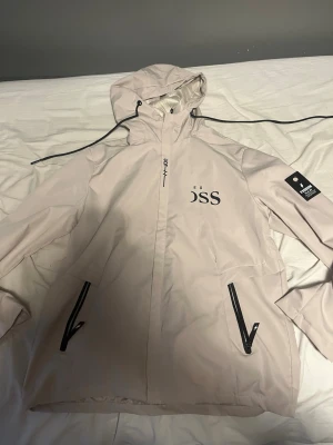 Beige vindjacka från Les Boss - Stilren beige vindjacka från Les Boss med svart logotyp på bröstet och ryggen. Jackan har huva med dragsko, svarta dragkedjor på fickorna och en patch på ena ärmen. Perfekt för blåsiga dagar och enkel att matcha med olika outfits.