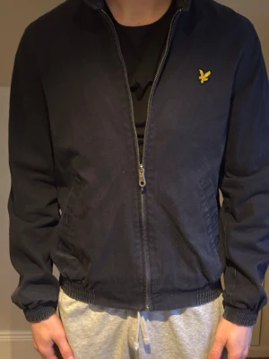 Marinblå bomberjacka från Lyle & Scott - Klassisk marinblå bomberjacka från Lyle & Scott med gul logga på bröstet. Jackan har dragkedja framtill, ribbade muddar vid ärmslut och midja samt två sidofickor. Insidan har ett rutigt foder för extra stilpoäng.
