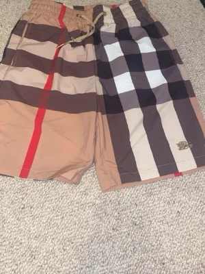 Burberry badbyxor med rutigt mönster - Snygga badbyxor från Burberry med klassiskt rutigt mönster i beige, brunt, svart, vitt och rött. De har resår och dragsko i midjan samt fickor på sidorna och en bakficka. Perfekta för stranden eller poolen. Materialet är snabbtorkande polyester.