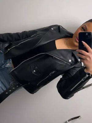 Svart skinnjacka med dragkedjor - Snygg svart skinnjacka i klassisk bikerstil med silvriga dragkedjor och detaljer. Jackan har bred krage, fickor med dragkedja och knappdetaljer. Perfekt för en cool och trendig look. Nästan helt oanvänd