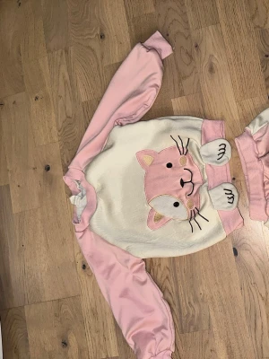 Söt set med kattmotiv i rosa - Söt set med kattmotiv i rosa Mysig tvådelad i mjuk fleece med långärmad tröja och byxor. Pyjamasen är ljusrosa och vit med ett stort broderat kattansikte på tröjan och ett mindre på byxan. Perfekt för dig som älskar gulliga djurmotiv och vill ha något bekvämt att chilla i.