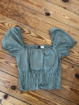 Grön croppad topp från H&M Divided - Trendig grön croppad topp från H&M Divided med puffiga korta ärmar och smockad midja. Tillverkad i mjuk bomull och har en fyrkantig ringning som ger en söt och modern look. Perfekt för dig som vill ha en stilren men ändå lekfull topp.