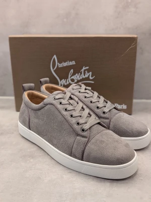 Gråa sneakers från Christian Louboutin - Stilrena gråa sneakers i mocka från Christian Louboutin med klassisk låg siluett, vita sulor och den ikoniska röda undersulan. Skorna har tonade skosnören och diskreta sömdetaljer. Perfekta för dig som vill ha en lyxig och trendig look.