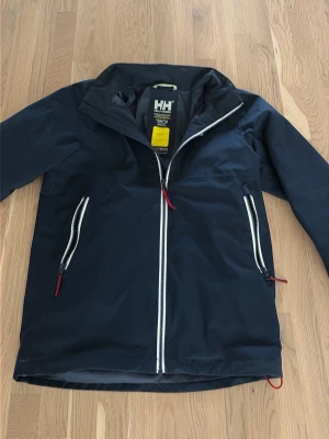 Svart vindjacka från Helly Hansen - Snygg svart vindjacka från Helly Hansen med vita dragkedjor och röda detaljer. Jackan har två sidofickor med dragkedja, hög krage och justerbar nederkant. Perfekt för blåsiga dagar och enkel att matcha med olika outfits. Det ända som är lite trasigt är fickorna inuti!