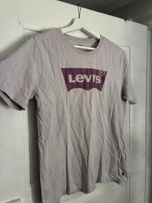 Beige/grå Levi's t-shirt med logga - Säljer en beige/grå t-shirt från Levi's med klassisk logga i lila och vitt framtill. T-shirten har rund halsringning och korta ärmar. Tillverkad i mjuk bomull, perfekt för en avslappnad stil.