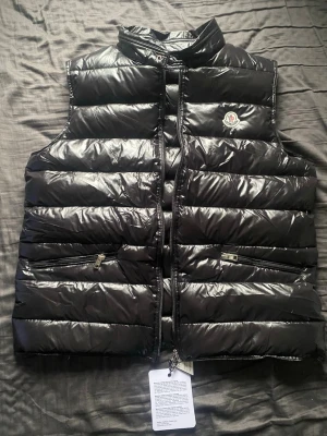 Svart dunväst från Moncler - Snygg svart dunväst från Moncler med glansig finish och klassisk logga på bröstet. Västen har ståkrage, två dragkedjefickor och quiltad design. Perfekt för lager-på-lager och streetstyle. Kan även passa L och M, beror på längd och bredd