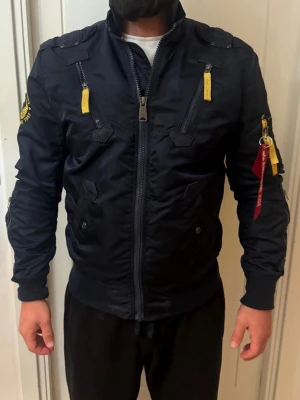 Mörkblå bomberjacka Alpha Industries - Snygg mörkblå bomberjacka från Alpha Industries med gula detaljer och dragkedjor framtill. Jackan har patch på ärmen, ribbade muddar och ikonisk röd tag på ärmfickan. U.S. Air Force-tryck i gult på ryggen. Tillverkad i slitstarkt syntetmaterial.