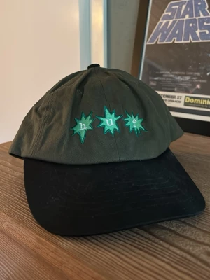 Grön och svart keps från HUF - Snygg keps från HUF i mörkgrönt tyg med svart ganska rak men lite böjd skärm. Framsidan har broderade gröna stjärnor med texten 'huf' i vitt. Justerbar snapback baktill för perfekt passform. Perfekt accessoar för en streetwear-look. Använd max 5 ggr då jag glömt bort den. Nypris 550