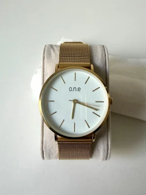 Guldig klocka från o.n.e med meshband - Stilren klocka från o.n.e med vit urtavla och guldiga detaljer. Klockan har ett elegant mesharmband i metall och minimalistisk design utan siffror. Perfekt accessoar för att lyfta din outfit 🤍