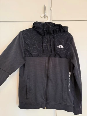 Svart vindjacka från The North Face - Snygg svart vindjacka från The North Face med mönstrad axelparti och huva. Jackan har dragkedja framtill, två fickor och justerbar huva med snörning. Perfekt för blåsiga dagar och har en sportig look med logga på bröstet. Priset kan diskuteras