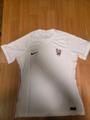Nike Frankrike slim fit Bortatröja Women's EURO 2022 Dam storlek L - Vit fotbollströja med korta ärmar. Tröjan har en broderad logotyp med en tupp och texten "FFF" på bröstet, samt en svart Nike-logotyp. Längs sidorna finns ett rosa mönstrat band. En etikett med texten "ENGINEERED Z699447YR0" sitter nedtill på framsidan. I nacken finns en tryckt etikett med Nike-logotyp, "DRI-FIT" och storleksinformation. En tygetikett med tvättråd och materialinformation finns också.