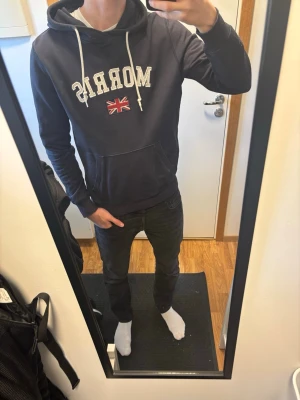 Morris mörkblå hoodie med tryck - Mörkblå hoodie från Morris med vit text och brittisk flagga på bröstet. Storlek M passar som S. Klassisk känguruficka och vita snören i huvan. Perfekt för dig som gillar stilren design och vill ha något bekvämt att slänga på dig. Materialet är mjukt och skönt.