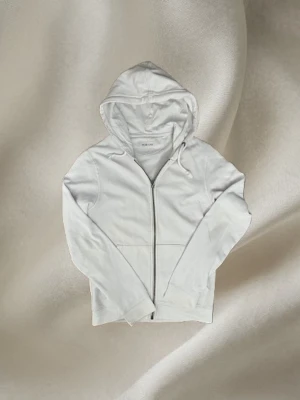 Vit hoodie med dragkedja från Pier One - Snygg vit hoodie från Pier One med dragkedja framtill, huva med dragsko och två fickor. Perfekt för en clean och stilren look. Tillverkad i mjukt material som känns skönt mot huden. Passar till det mesta och är enkel att matcha. Xs