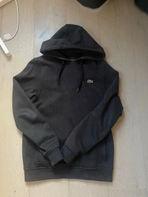 Svart hoodie från Lacoste - Svart hoodie från Lacoste med klassisk krokodillogga på bröstet. Tröjan har huva med dragsko och ribbade muddar vid ärmslut och nederkant. Perfekt för en chill och stilren look.