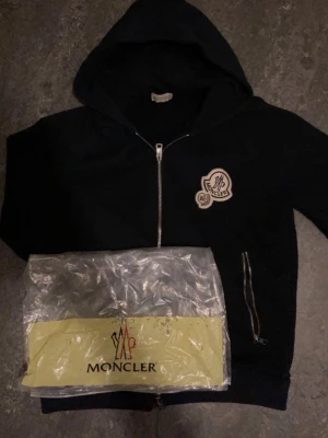 Mörkblå hoodie med dragkedja från Moncler - Hej, säljer en fet model på luvtröjor från moncler, speciell design med det stora märket. Den har nfc skan och är o fint skick. Den ända nackdelen är att dragkedjan är lite trög, annars är det ett skön tröja nu inför sommaren. Passa på och skriv vid fundering