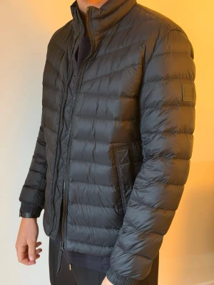 Svart quiltad pufferjacka från BOSS - Snygg svart pufferjacka från BOSS med quiltad design och röd insida. Ny skick, storlek medium. 