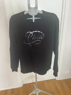 Svart sweatshirt från Philipp Plein - Cool svart sweatshirt från Philipp Plein med glansigt logotryck på bröstet. Tröjan har rund halsringning och långa ärmar. Perfekt för en avslappnad och trendig stil.