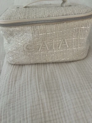 Vit necessär från CAIA i tweed - Säljer en snygg vit necessär från CAIA i glittrig tweed med dragkedja och handtag upptill. Perfekt för smink eller småprylar. Stilig design med CAIA-logga broderad på framsidan och silvriga detaljer.