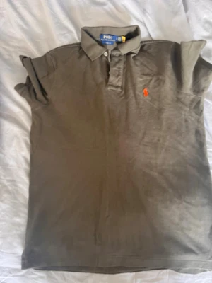 Olivgrön pikétröja från Polo Ralph Lauren - Snygg olivgrön pikétröja från Polo Ralph Lauren med klassisk krage, korta ärmar och orange broderad logga på bröstet. Tillverkad i mjuk bomull som känns skön mot huden. Perfekt till jeans eller shorts för en avslappnad look.