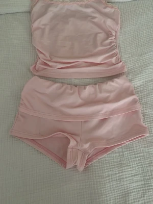Ljusrosa linne och shorts set - Sött set med linne och shorts i ljusrosa färg. Linnet har tunna axelband och spetsdetaljer upptill, medan shortsen har en mjuk och bekväm passform. Perfekt för att chilla hemma eller som mysigt nattlinne. Materialet känns mjukt mot huden.