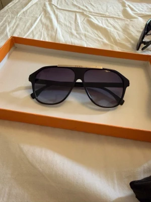 Svarta Fendi pilotbågar solglasögon - Snygga svarta solglasögon från Fendi med klassisk pilotform och mörka glas. Bågarna har en metallist upptill med Fendi-loggan och breda skalmar. Perfekta för att lyfta din stil under soliga dagar.
