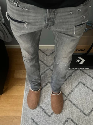 Grå Jack & Jones jeans slim - Säljer ett par grå slim jeans med slitningar och trasiga detaljer framtill. Jeansen har klassisk femficksdesign och dragkedja. Snyggt tvättade med en modern, lite ruffig look. Perfekta för dig som gillar streetstyle.