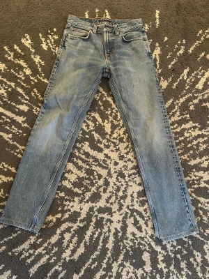 Blå raka jeans från Nudie Jeans - Säljer ett par klassiska blå raka jeans från Nudie Jeans med fem fickor och snygga kontrastsömmar. Jeansen har en ljus tvätt och ikoniska broderade bakfickor. Perfekta för en avslappnad och trendig stil.