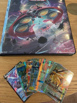 Pokémonkort med samlarpärm - Samling av Pokémonkort som sonen säljer med holografiska motiv och en färgglad samlarpärm med tryck. Korten har glittriga ytor och motiv i flera färger, bland annat blå, gul och röd. Perfekt för dig som gillar Pokémon och samlarkort.