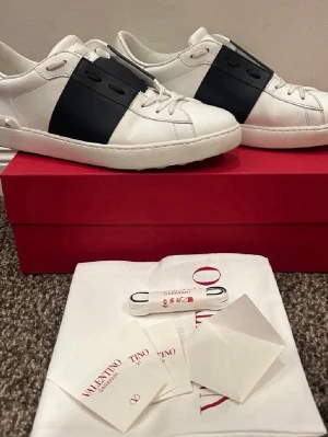 Valentino skor - Snygga vita sneakers från Valentino med en bred svart läderdetalj över mitten. Klassisk låg modell med rund tå och vita snören. Skorna har diskret logga på plösen och kommer med originalkartong och tillbehör. Perfekta för dig som gillar stilrena och exklusiva sneakers.