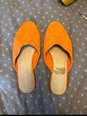 Orange tofflor från Attitude - Snygga orangea tofflor från Attitude med öppen häl och spetsig tå. Ovandelen är i mockaliknande material och sulan är i gummi. Perfekta för dig som vill sticka ut med färg och stil.