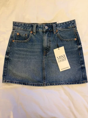 Blå jeanskjol mini från Stradivarius - Trendig blå jeanskjol i minilängd från Stradivarius. Klassisk femficksmodell med bälteshällor och knappstängning framtill. Perfekt för att skapa en cool och avslappnad look. Gjord i slitstarkt denim-material. Helt oanvänd 