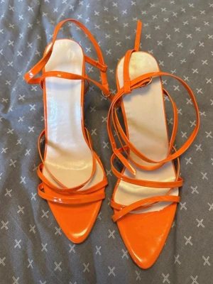 Orange lackpumps med remmar - Snygga orange pumps med smala remmar och spetsig tå. Skorna har höga klackar och är gjorda i glansigt lackmaterial. Perfekta för dig som vill sticka ut med färg och trendig design.