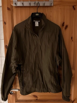 Olivgrön vindjacka från hope  - Snygg olivgrön vindjacka från hope  med halv dragkedja framtill och hög krage. Tillverkad i lätt, prassligt material som ger en avslappnad look. Jackan har resår vid ärmslut och nederkant samt två sidofickor med dragkedja.
