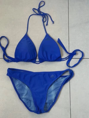Blå bikini från Adidas med knyt i storlek 40, endast provad - Snygg blå bikini från Adidas i storlek 40med klassisk trekantsmodell på överdelen och knyt i nacken och ryggen. Bikinin är endast provad. Underdelen är enkel och sportig med Adidas-logga på båda delarna. Perfekt för stranden eller poolen.