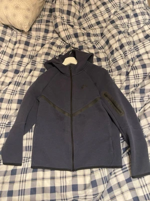 Nike Tech Fleece hoodie i mörkblå - Nike Tech Fleece hoodie i mörkblått med hel dragkedja och huva. Snygg detalj med svart diagonal linje över bröstet och en praktisk ficka med dragkedja på ärmen. Perfekt för en sportig och stilren look.