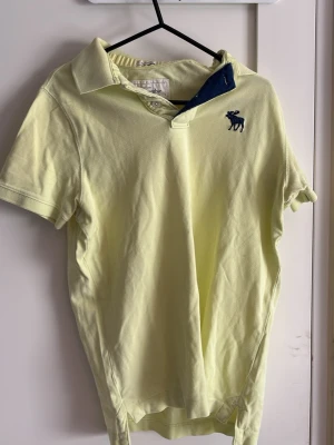 Gul pikétröja från Abercrombie - Ljusgul pikétröja från Abercrombie & Fitch med klassisk krage och korta ärmar. Tröjan har blå broderad älg-logga på bröstet och blå detalj på insidan av kragen. Perfekt för en avslappnad och stilren look.